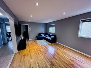 A modern living room with a dark grey wall, a wooden floor, a blue sectional sofa, and a flat-screen TV mounted on the wall. - Family Comfort Retreat (Brampton)