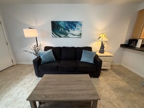 A living room with a black sofa, a wooden coffee table, a painting of a wave, a lamp, and a small plant.