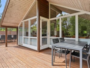 Eine überdachte Terrasse mit Tisch und Stühlen, großen Fenstern und einer Holzbalkendecke.