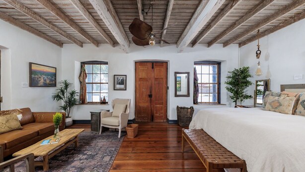 A room with a wooden ceiling, a large bed, a sofa, a chair, a small table, and a window. - Historic Rumpel Kammer - Steps to Main Street (Fredericksburg)