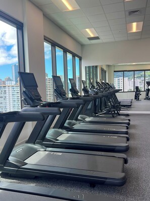 A row of stationary treadmills in a gym with large windows and a city view. - Newly Built High-Floor Condo with Diamond head & Ocean Views & Luxury Amenities (Honolulu)