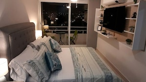 Een slaapkamer met een bed, een televisie, een plafondventilator en een balkon met uitzicht op de stad.