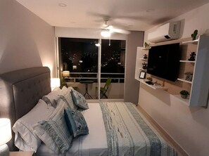 Een slaapkamer met een bed, een televisie, een plafondventilator en een balkon met uitzicht op de stad.
