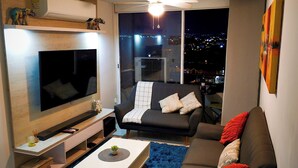 Een moderne woonkamer met een flatscreen televisie, een bank, een salontafel en uitzicht op de stad 's nachts.