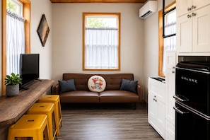 Un espacio de sala acogedor con un sofá marrón, una mesa de madera con un televisor, taburetes amarillos, una ventana con persianas y una zona de cocina con un electrodoméstico negro.