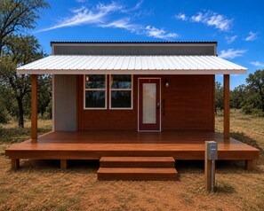 A small, single-story wooden cabin with a metal roof, a front door, and a set of steps leading to the entrance. - Live Oak Cabin: A Modern Escape at the Starlight PK Ranch on Possum Kingdom Lake (Graham)