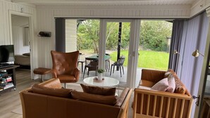 A living room with a brown leather sofa, a wooden chair, a TV stand, and a sliding glass door leading to an outdoor patio. - 4 Star Holiday Home in Esbjerg V (Esbjerg)