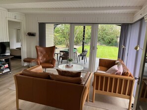 A living room with a brown leather sofa, a wooden chair, a TV stand, and a sliding glass door leading to an outdoor patio. - 4 Star Holiday Home in Esbjerg V (Esbjerg)