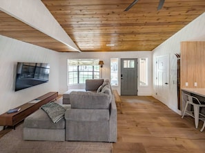 A living room with a grey sofa, a flat-screen TV, a wooden ceiling, and a large window. - Bear Crest W/Spa Near Snow Summit (Big Bear Lake)