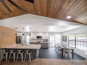A modern kitchen with a central island, bar stools, a dining area with a table and chairs, and a view through large windows. - Bear Crest W/Spa Near Snow Summit (Big Bear Lake)