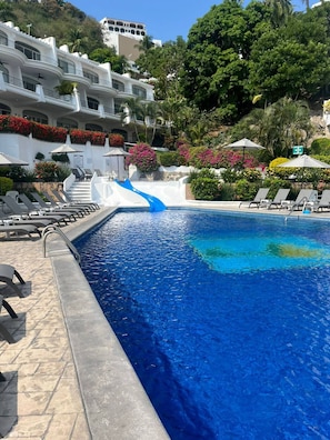 Ein Schwimmbecken mit einer blauen Rutsche, Liegestühlen und einem weißen Gebäude mit Balkonen.
