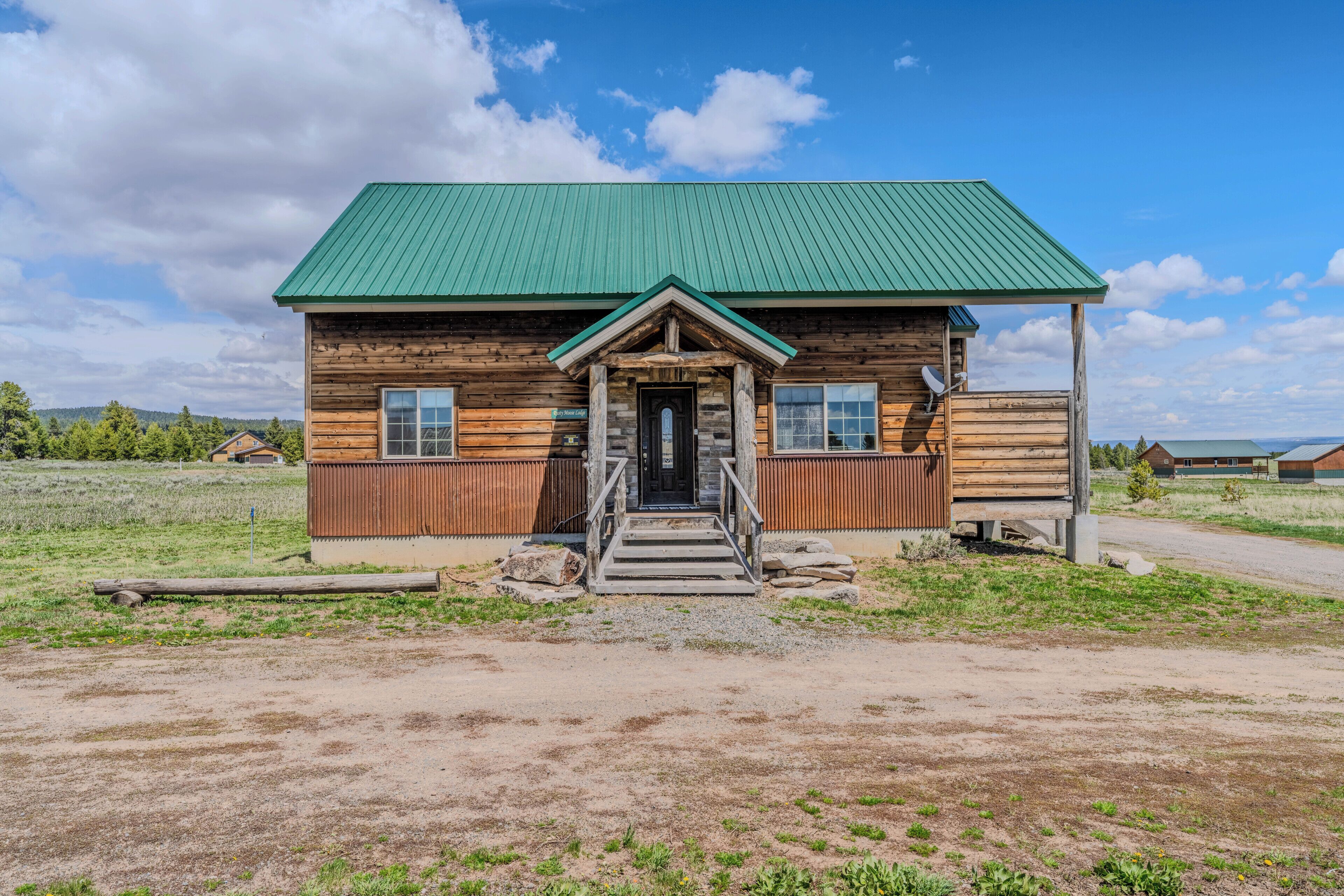 A wooden cabin with a green roof, a front porch, and a dirt driveway.
