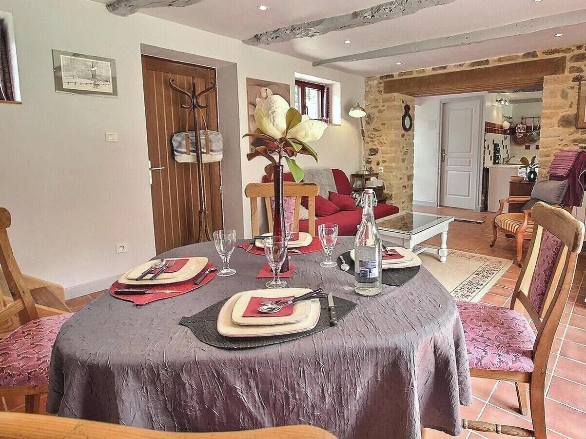 Un comedor con una mesa redonda dispuesta para cuatro, un jarrón con una flor, y una cocina visible al fondo.