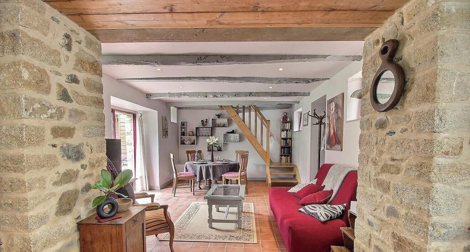 A rustic interior with exposed beams, stone walls, a red sofa, a dining area, and a staircase.