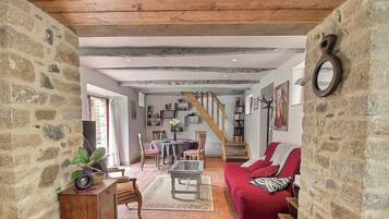 A rustic interior with exposed beams, stone walls, a red sofa, a dining area, and a staircase.