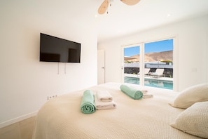 A bedroom with a bed, a flat-screen TV, a sliding glass door to a pool area, and a ceiling fan. - Casa Luna by BLVE Homes (Yaiza)