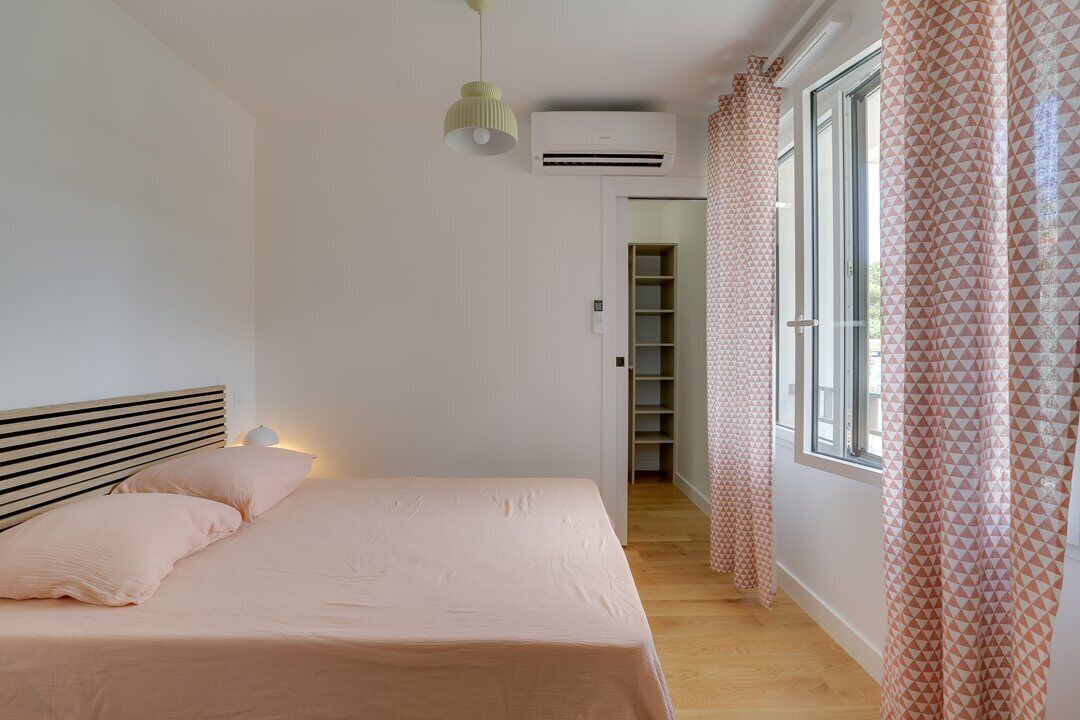A bedroom with a bed, a bedside lamp, an air conditioning unit, a window with curtains, and a hallway leading to a closet.