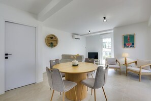 Ein modernes Wohnzimmer mit einem runden Holztisch, grau gepolsterten Sesseln, einer Couch, einem Fernseher und einer wandmontierten Klimaanlage.