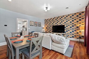 A modern living room with a sofa, TV, and dining table set with orange napkins. - Trendy 2BR Near Carson Street | Free Parking (Pittsburgh)
