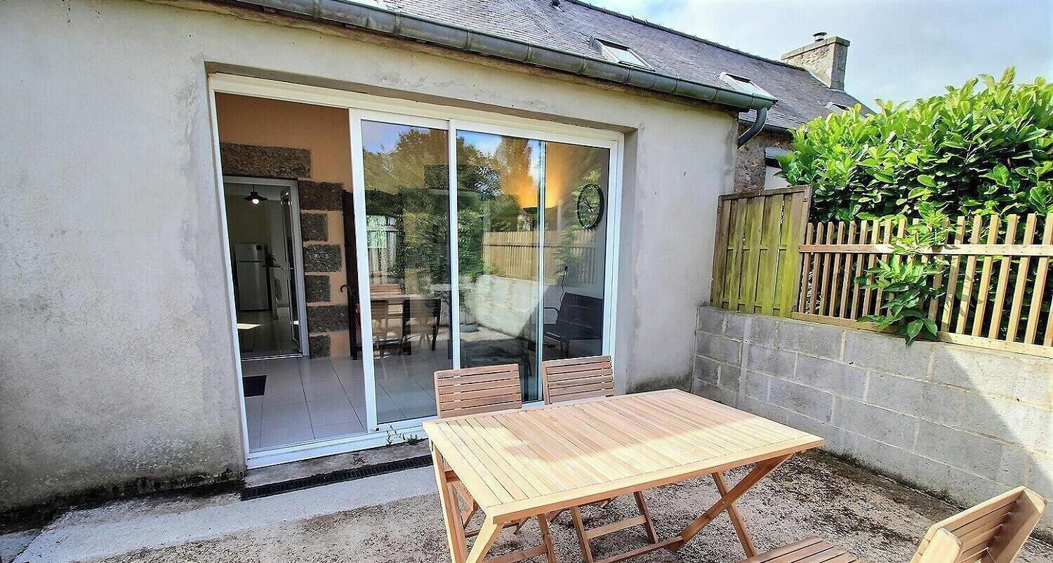 A patio with a wooden table and chairs, a sliding glass door leading to an interior space, and a wooden fence with greenery.