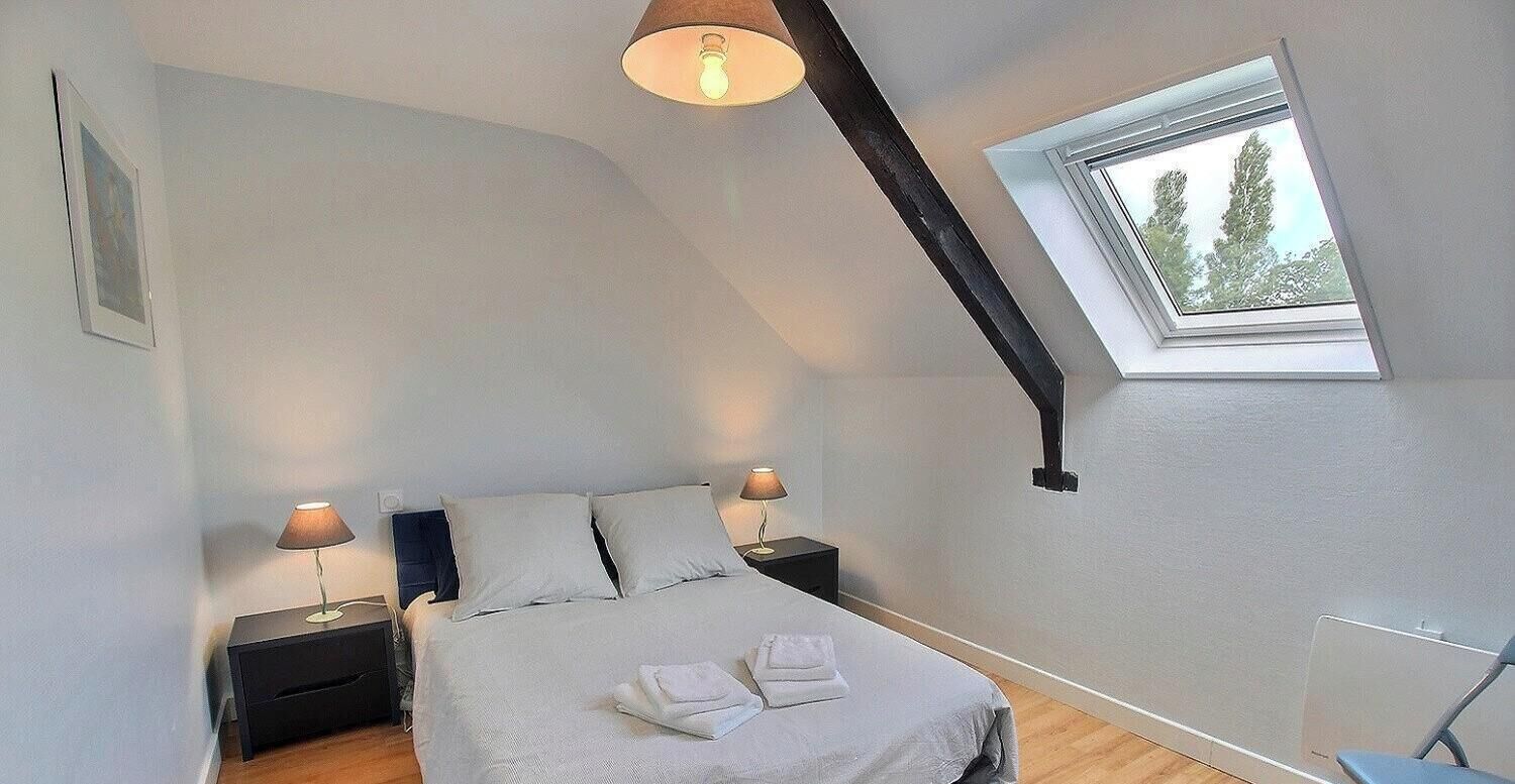 Quarto com teto inclinado, uma cama com almofadas, duas mesas de cabeceira com luminárias e uma janela com vista para árvores.