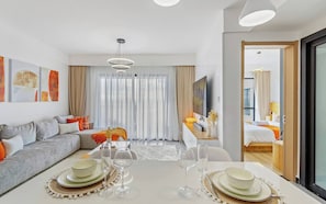 Ein modernes Hotelzimmer mit Essbereich, einer Couch, einem Fernseher und einem großen Fenster mit Vorhängen.