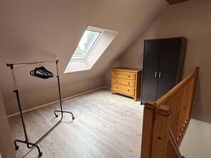 Ein Zimmer mit schrägverdeckter Decke, einem Oberlicht, einem hölzernen Schminktisch, einem Kleiderschrank und einem Metallgestell mit Kleiderbügeln.