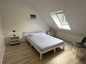 Ein Schlafzimmer mit schrägen Wänden, einem Oberlicht, einem Bett, einem Sessel und einem Nachttisch mit Lampe.