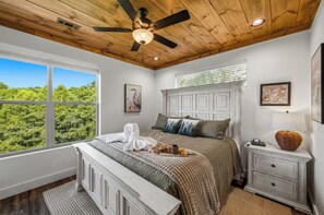 Ein Schlafzimmer mit Holzbalkendecke, einem großen Fenster, einem Bett mit Decke und Kissen, einem Nachttisch mit Lampe und einem gerahmten Bild an der Wand.