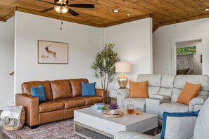 Un salon avec un canapé en cuir marron, une table basse blanche et un fauteuil gris. On y trouve un ventilateur de plafond, un tableau encadré au mur et une plante en pot.