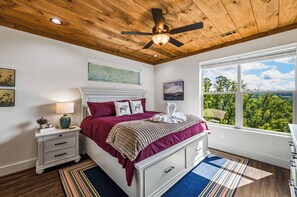 A bedroom with a wooden ceiling, a bed with a red bedspread, a nightstand with a lamp, and a window with a view of trees. - Smokey Mountain High (Gatlinburg)