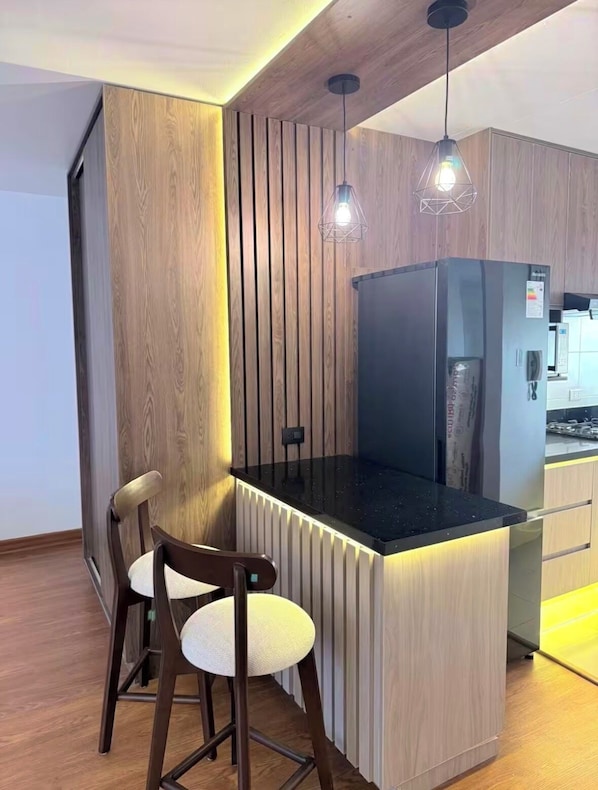 A modern kitchen with a wooden accent wall, a black countertop island, two pendant lights, and a refrigerator. - Modern, Central and Cozy | San Isidro (San Isidro)