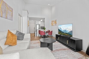 A modern living room with a sofa, coffee table, and TV. There is a kitchen in the background with red chairs. - 2302 Cortland (Houston)