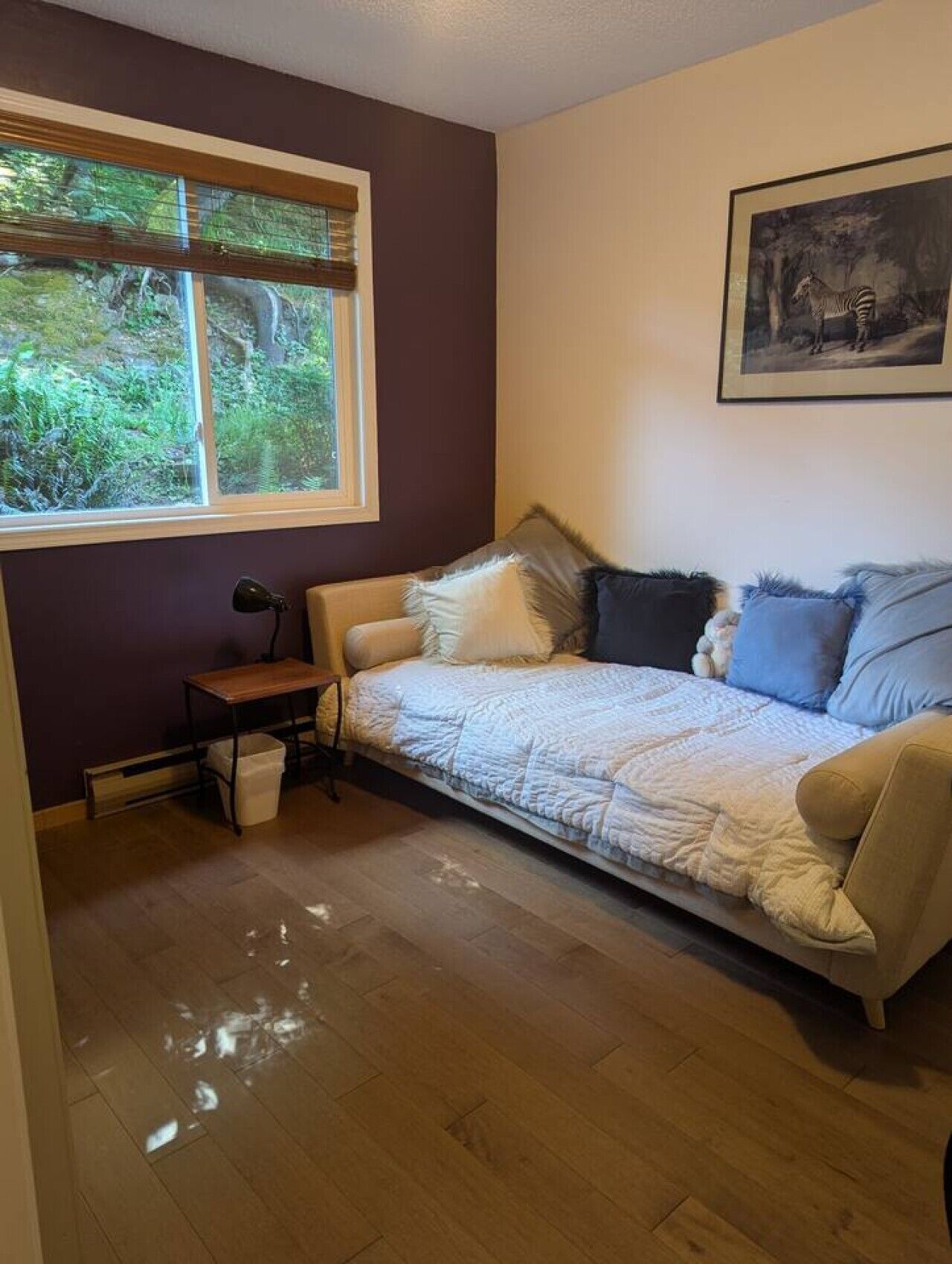 Ein Zimmer mit einer Couch, einem kleinen Tisch, einer Lampe, einem gerahmten Bild an der Wand und einem Fenster mit Blick auf Grünflächen.