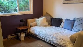 Ein Zimmer mit einer Couch, einem kleinen Tisch, einer Lampe, einem gerahmten Bild an der Wand und einem Fenster mit Blick auf Grünflächen.