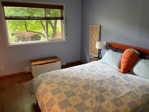 Une chambre à coucher comprenant un lit, une table de chevet, une lampe, une fenêtre avec des stores et une vue sur les arbres.