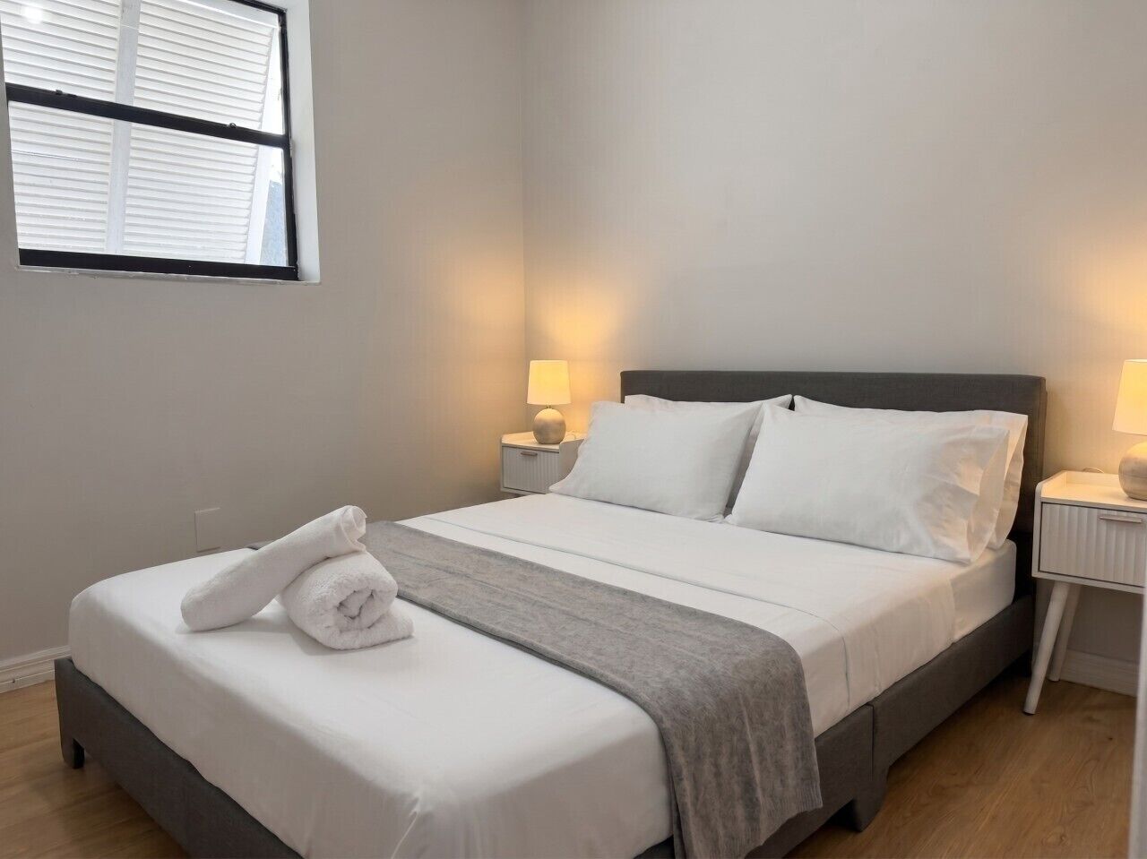 Una cama bien hecha con sábanas blancas, un plaid gris y una toalla enrollada en el pie. Hay dos lámparas de noche en mesitas blancas.