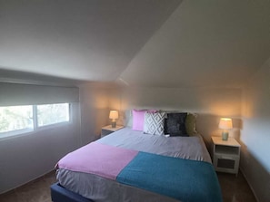 Ein Schlafzimmer mit einem Bett, zwei Nachttischen, einem Fenster und einem Beistelltischchen mit einer Lampe.