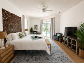 Ein Schlafzimmer mit Bett, Fernseher, Deckenventilator, Fenster mit Vorhängen und Blick ins Freie.