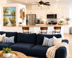 Ein Wohnzimmer mit einem dunkelblauen Sofa, drei Sesseln, einem Couchtisch mit Pflanze und Kerze sowie einem Strandbild. Zusätzlich ist eine Küche mit weißen Schränken, einem Kühlschrank und einer Mikrowelle zu sehen.