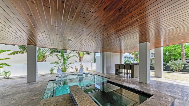 Uma área de piscina moderna ao ar livre, com teto de madeira, piso de pedra e piscina com fundo de vidro.