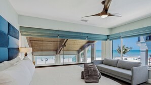A hotel room with a large bed, a sofa, a ceiling fan, and a view of the beach.