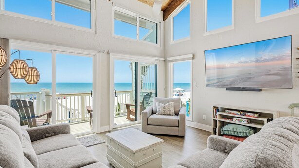 A spacious living room with a large TV, comfortable seating, and a view of the ocean through large windows.