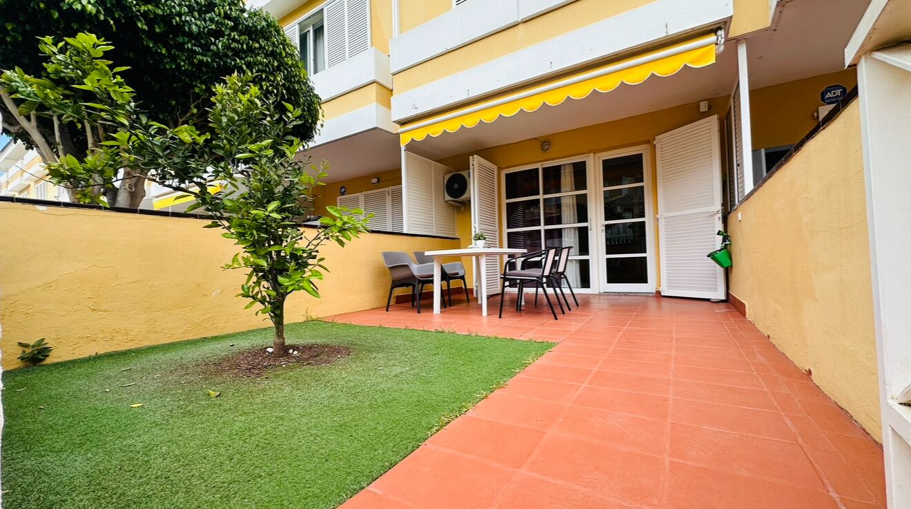 Un patio con un pequeño césped, una mesa con sillas y un edificio con toldo amarillo.