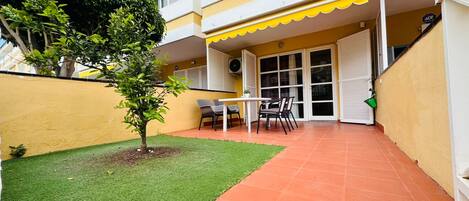 Un patio con un pequeño césped, una mesa con sillas y un edificio con toldo amarillo.