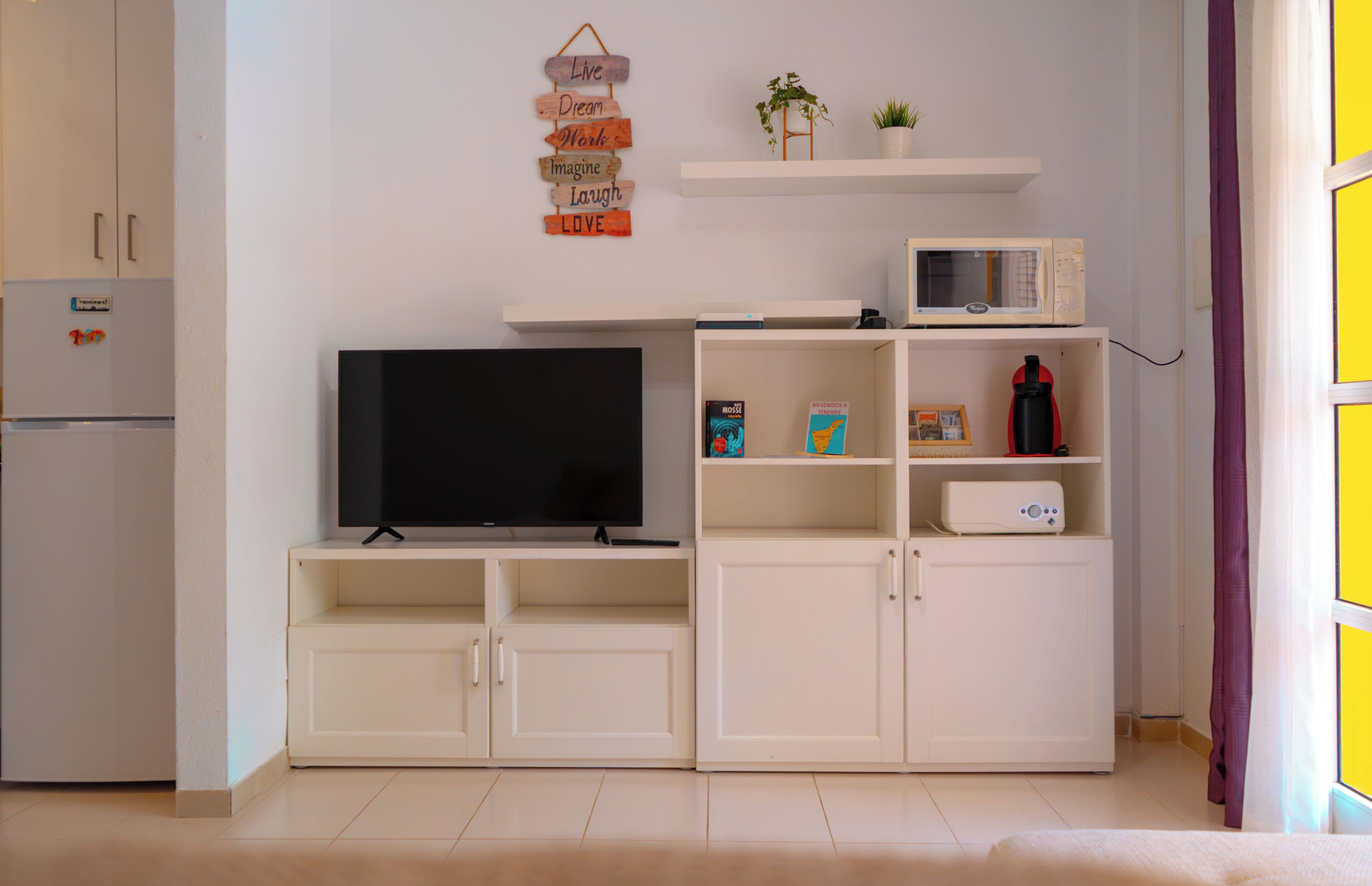 A modern kitchen with a flat-screen TV, white cabinetry, a microwave, and a small plant on a shelf.