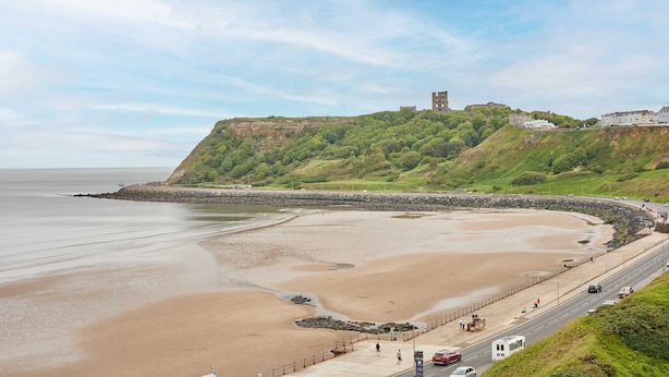 Une route côtière bordée d’une plage, un château perché sur une colline et une route menant à un bâtiment.