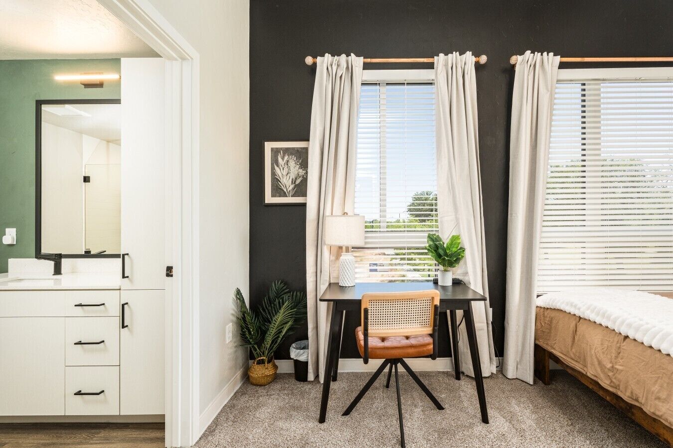 Een slaapkamer met een bed, een bureau met stoel, een raam met gordijnen, een nachtkastje en een wandlamp.