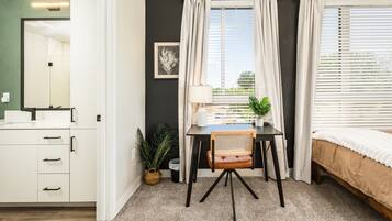 Een slaapkamer met een bed, een bureau met stoel, een raam met gordijnen, een nachtkastje en een wandlamp.