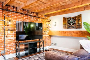 Un salon avec un mur en briques, un téléviseur à écran plat, un jeu de fléchettes et un coin salon confortable.